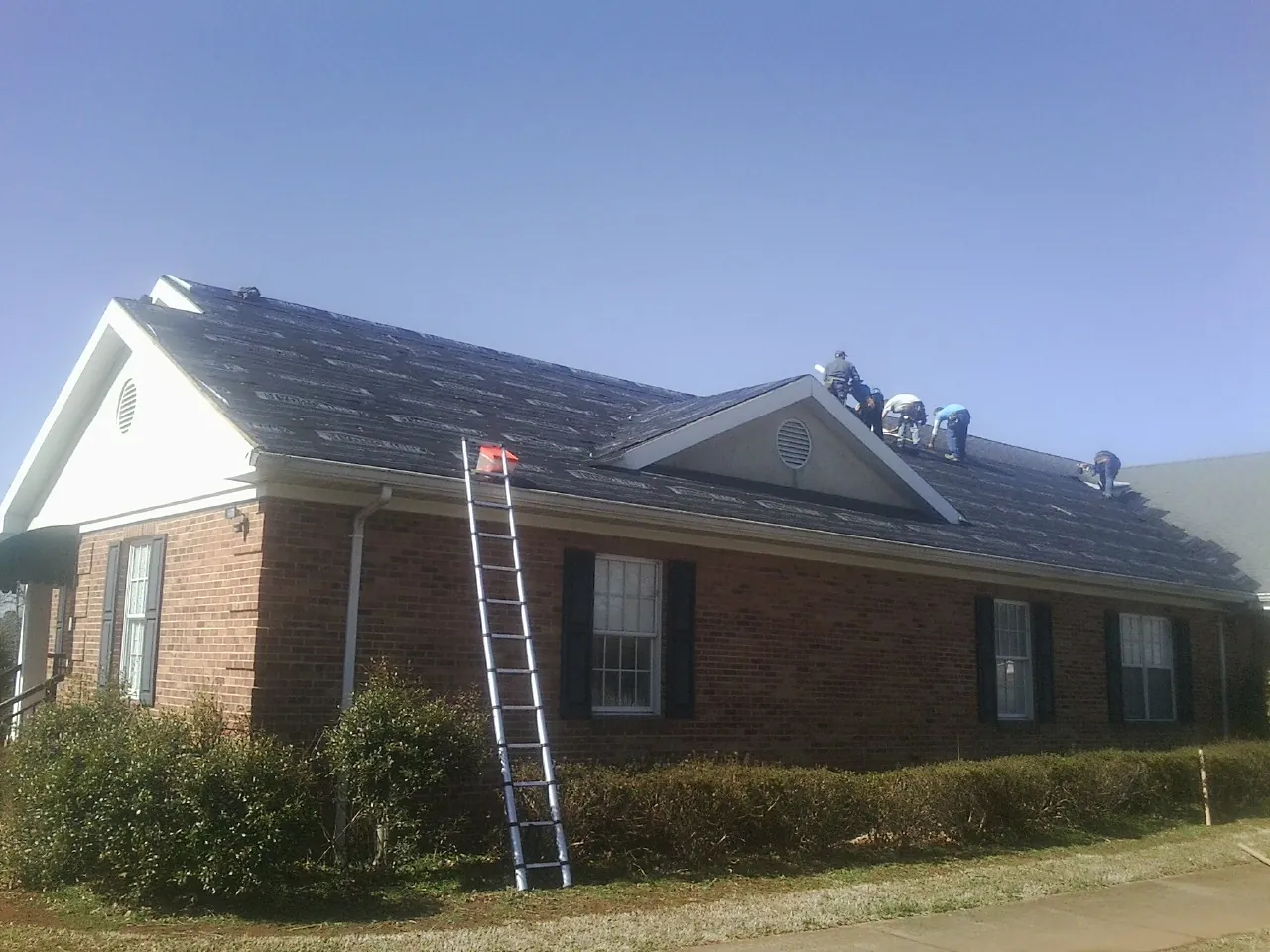Crew tearing shingles off a brick ranch roof with a ladder set — Emergency Roof Repair work by our crew in Triangle
