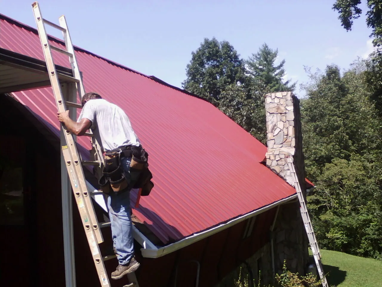 Roofer on a ladder beside a red metal roof and stone chimney — Soffit & Fascia Repair by Roofing Brothers in Triangle